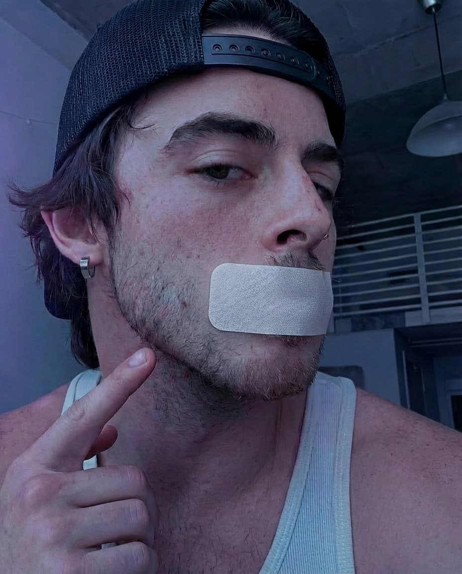 Man with bandage over mouth pointing to chin in indoor setting